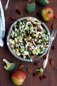 Healthy Broccoli Salad with Vegan Bacon, Apples, Blue Cheese and Pecans Pouring Dressing Overhead on the Bowl with the Ingredients Around