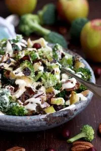Healthy Broccoli Salad with Vegan Bacon, Apples, Blue Cheese and Pecans Pouring Dressing Closeup on the Dish with Apples Blurred in the Background