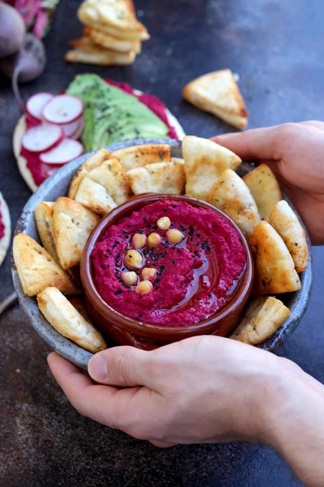 Hands Holding a Dish with Beet Hummus Surrounded by Pita Chips