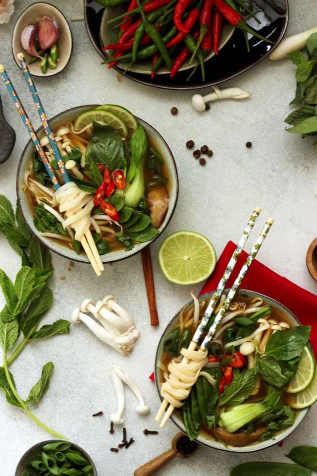 Two Bowls of Vegan Pho Soup with Plenty of Ingredients Around