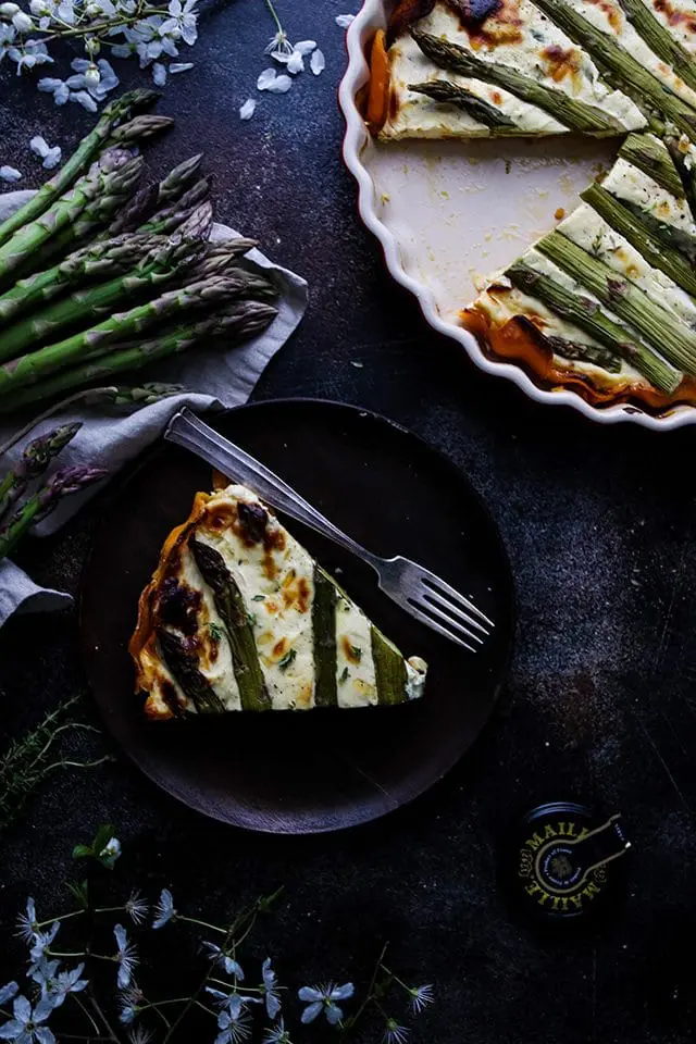 A Slice of Quiche on a Wooden Plate Next to a Pie Pan and Fresh Asparagus Spears