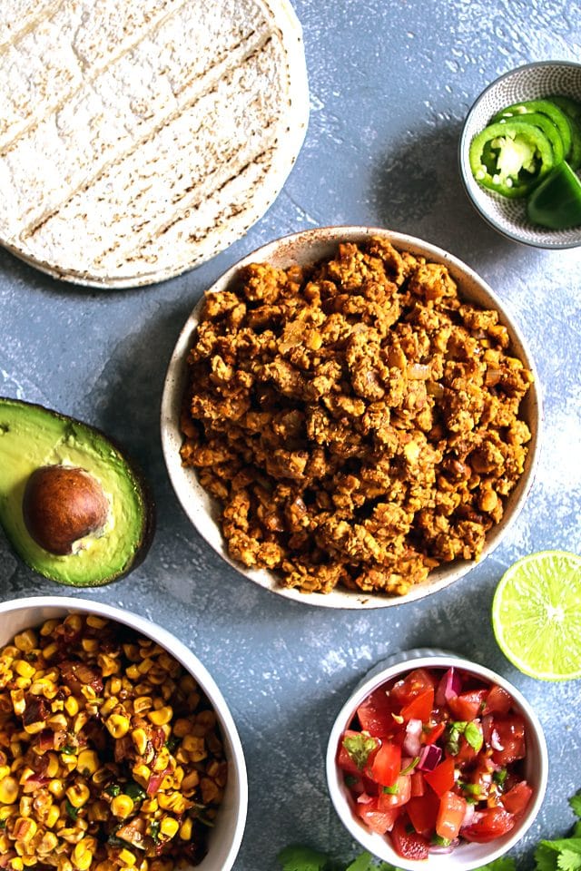 Tempeh Taco Meat on a Plate Surrounded by Tortillas, Jalapenos, Corn Salsa, Avocado, Pico de Gallo and Lime