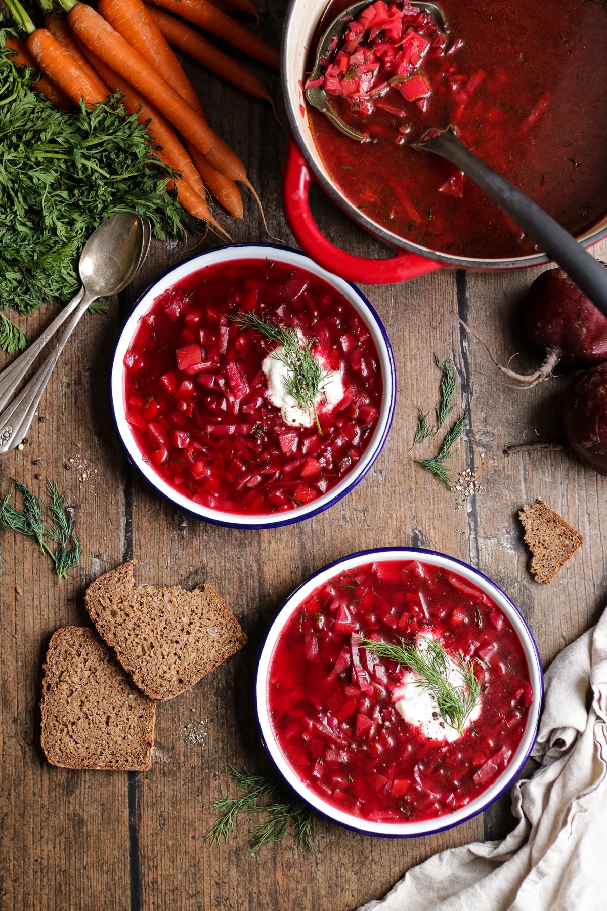 Two plates with borscht next to a pot and carrots surrounded with sliced rye bread.