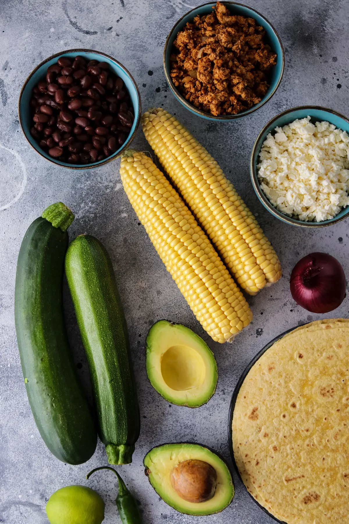 Ingredients for grilled tacos including zucchini, corn, avocado, tempeh taco meat, feta cheese, black beans, tortillas, etc.